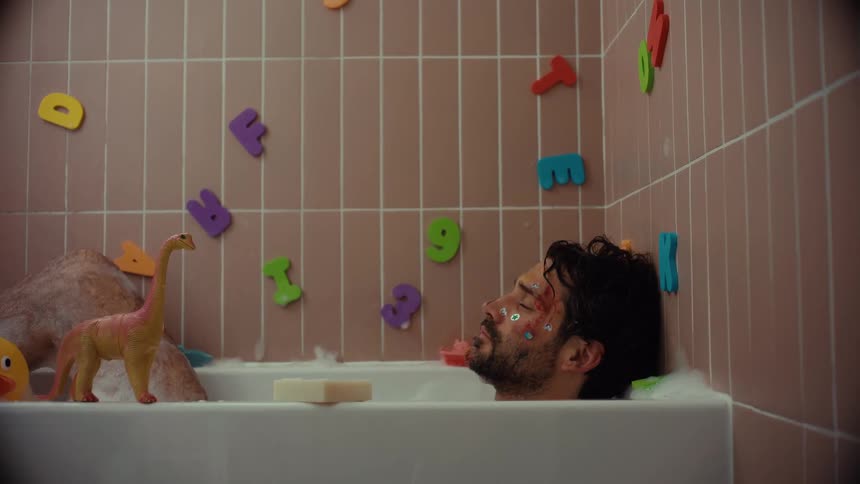 An exhausted man covered in stickers and paint falls asleep in a bubble bath surrounded by children's toys
