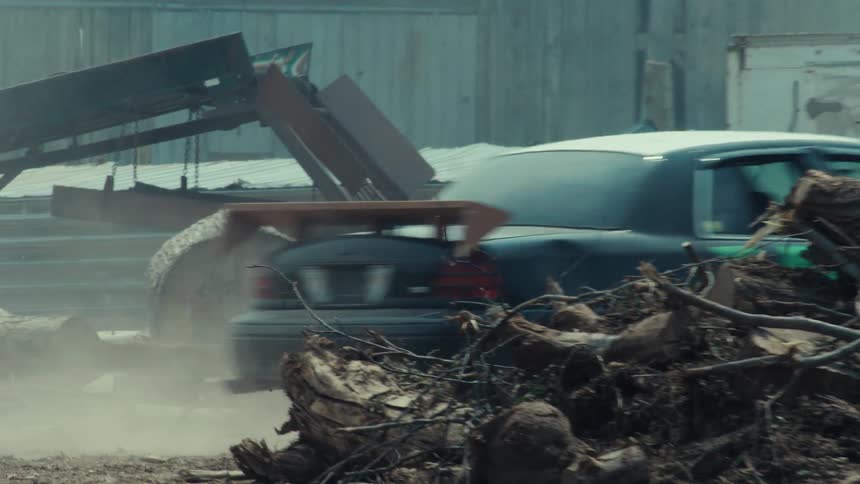 A graffiti-covered car drives in slow motion through a dusty industrial yard past a pile of debris