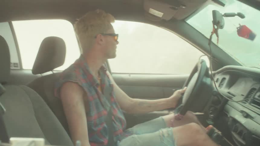 A man in a sleeveless plaid shirt drives a car through a hazy, dusty area and then picks up a paintball gun