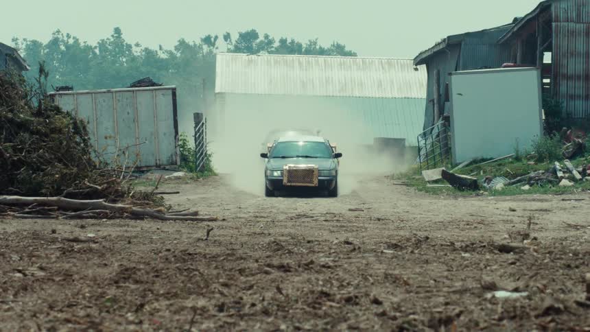 Two demolition derby cars race down a dusty dirt road toward the camera in slow motion