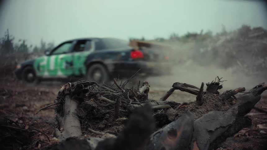 A black sedan with green graffiti drives through a desolate, burnt wasteland, kicking up dust and smoke