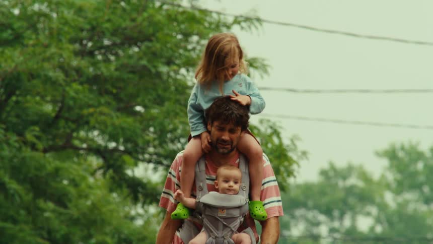 A father expertly multitasks, carrying his baby and toddler while pushing a stroller and holding a bike on a neighborhood walk