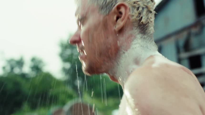 A rugged tattooed man with bleached blonde hair enjoys a refreshing outdoor shower, washing his hair in slow motion
