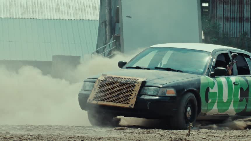 A modified car does a burnout in slow motion, kicking up a cloud of dust as a passenger aims a paintball gun out the window