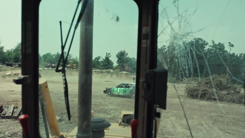 From the cracked windshield of a bulldozer, a race car with green lettering speeds through a dusty industrial junkyard