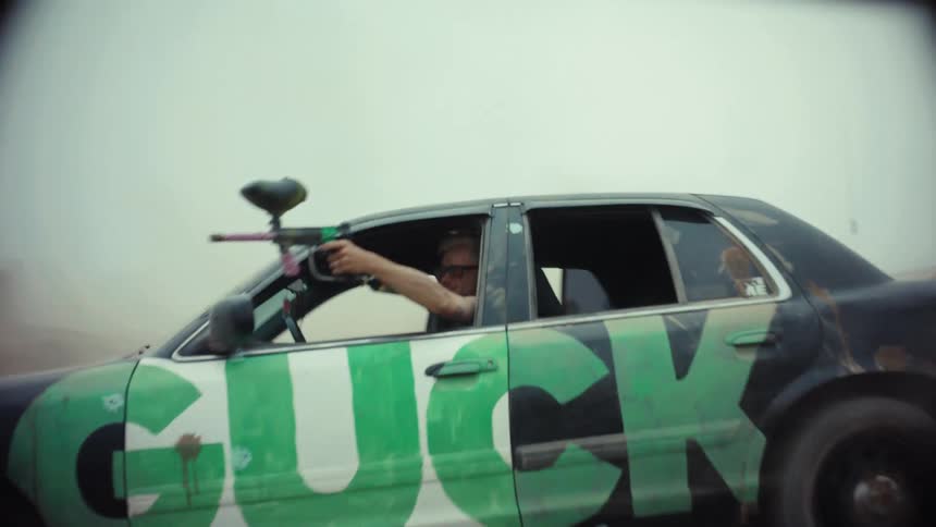 A man in a graffiti-covered car shoots a paintball gun while speeding down a dirt road, kicking up a large cloud of dust in slow motion