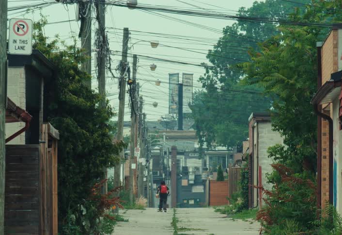 A person with a red backpack skateboards away from the camera down a long, overgrown urban alley on an overcast day