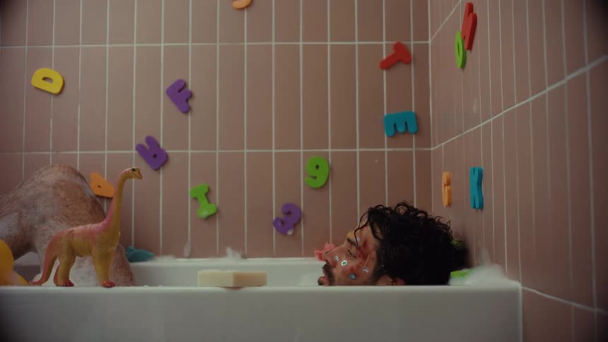 A bruised and battered man with glitter on his face sinks into a bubble bath surrounded by children's toys