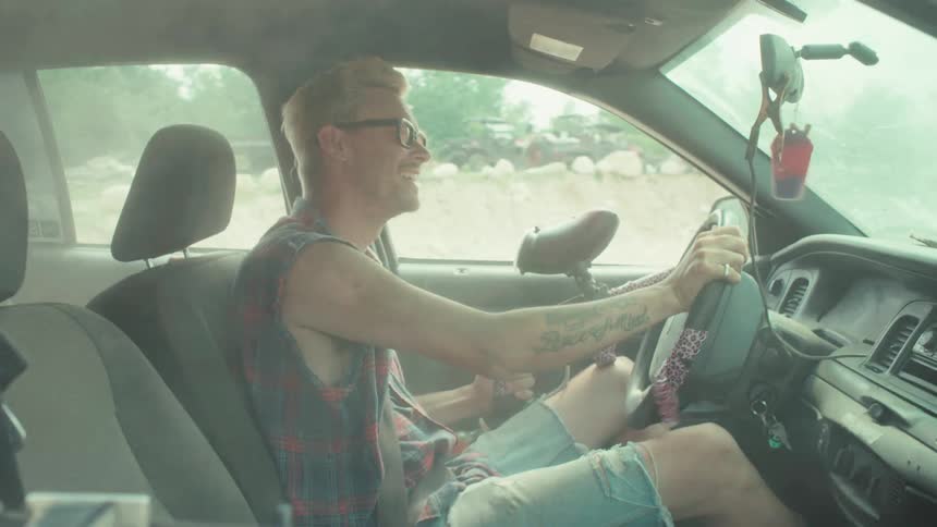 A man with a paintball gun joyfully drives a car through a dusty and smoky landscape in slow motion