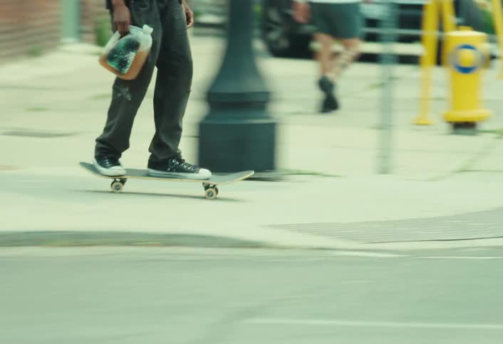 Tracking shot of a young man with a red backpack skateboarding along a city sidewalk with traffic in the foreground