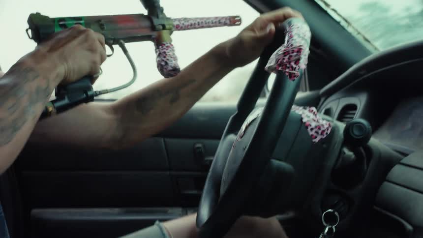 A wild-eyed man with a paintball gun laughs maniacally while chaotically driving a car through a rural, post-apocalyptic landscape