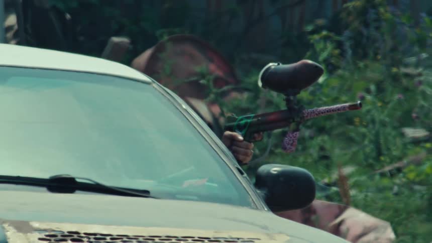 A paintball player slowly takes aim with their marker from behind a white car in an overgrown field