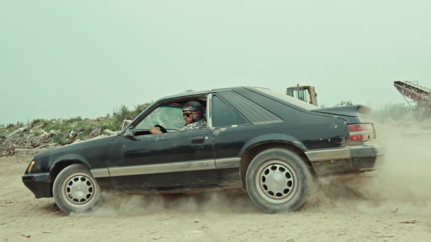 A man in a classic black sports car performs a dramatic burnout on a dusty road in slow motion, kicking up a huge cloud of dust as he drives off