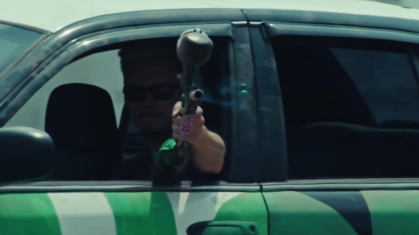 A paintball player shoots from the window of a moving camouflage car in extreme slow motion