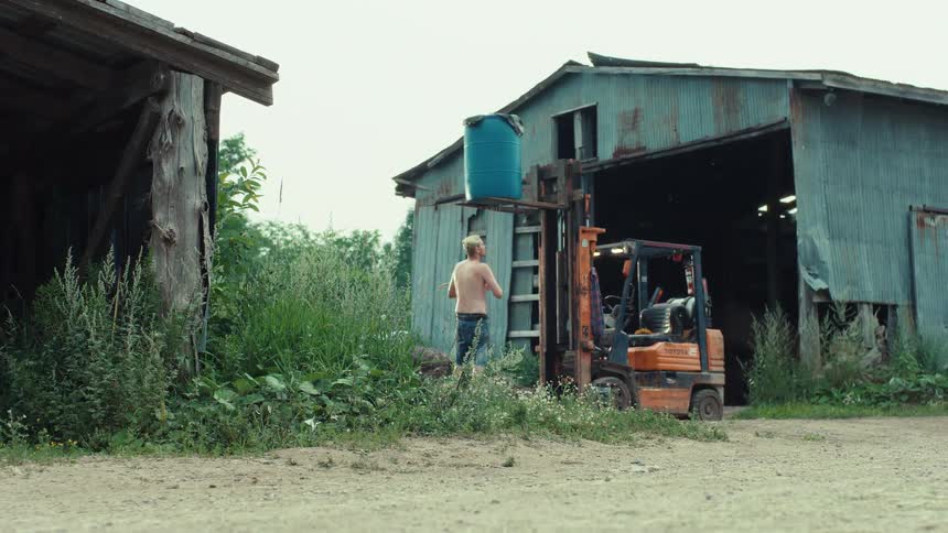 A shirtless man stretches while working with a forklift to lift a blue barrel next to a rustic metal barn