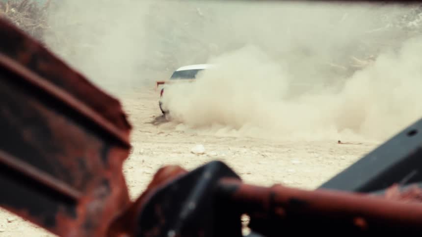 A modified race car drifts through a dusty junkyard, kicking up a massive cloud of dirt in slow motion