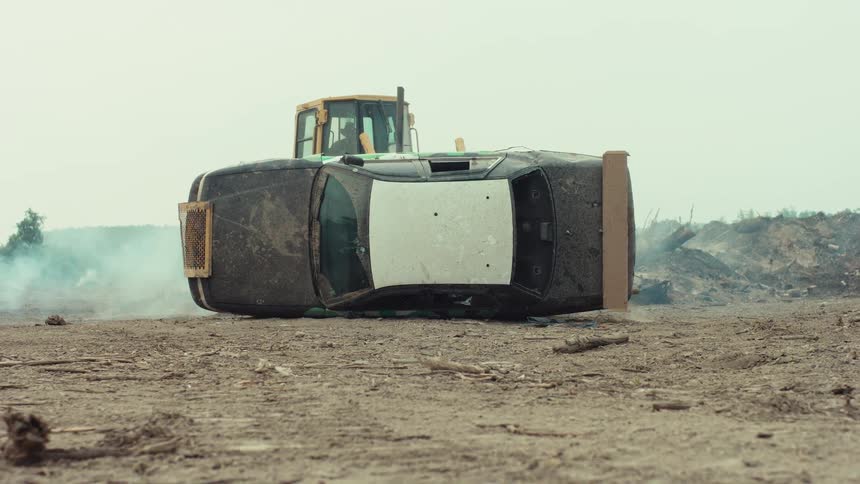 A powerful yellow bulldozer slowly flips a wrecked black car onto its roof in a dusty, barren junkyard