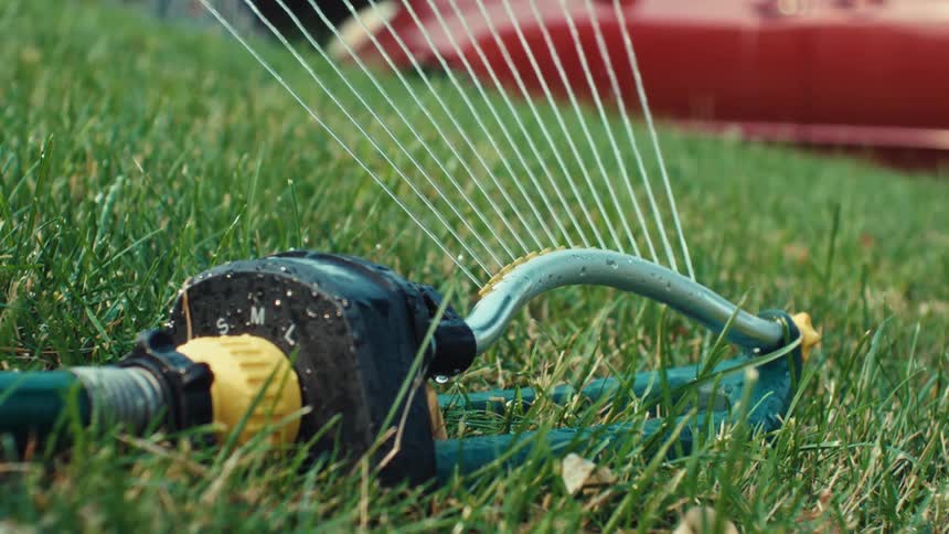 A low-angle close-up shot shows an oscillating lawn sprinkler watering a lush green lawn on a sunny day