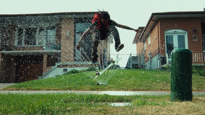 A stylish skateboarder performs a kickflip in super slow motion through a lawn sprinkler's spray in a suburban neighborhood