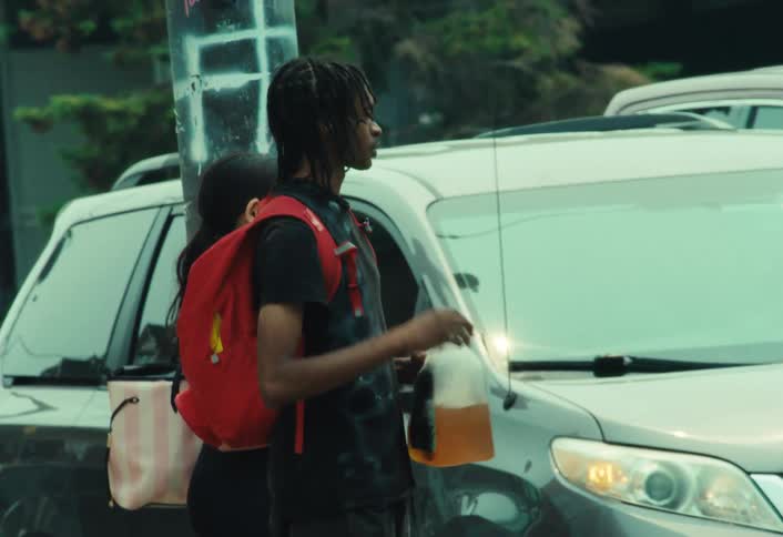 A young man with a skateboard takes a refreshing drink from a large jug while waiting on a busy city street corner
