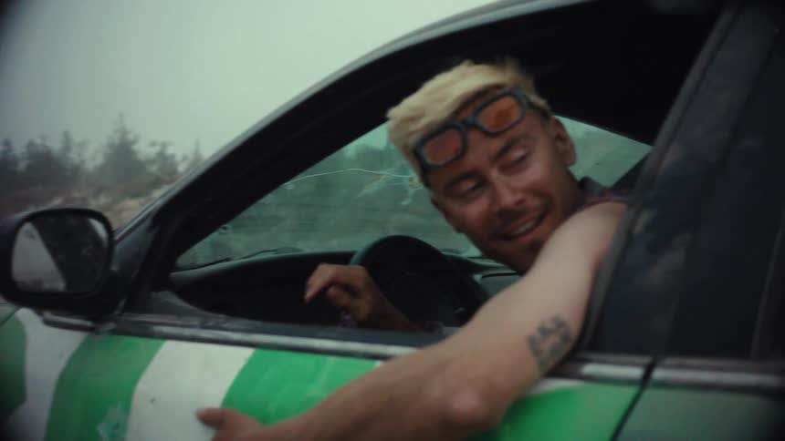 A smiling man with blonde hair gives a thumbs-up from his graffiti-covered car before speeding off and kicking up dust