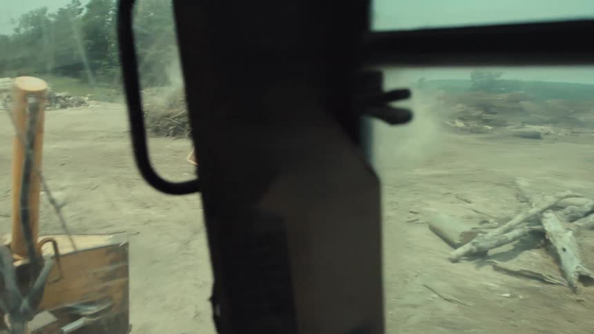 A modified race car kicks up dust as it speeds by in a lumberyard, seen from the perspective of a bulldozer operator