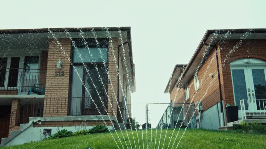 A slow-motion, tilt-up shot captures a water sprinkler nourishing a lush green front lawn in a suburban setting
