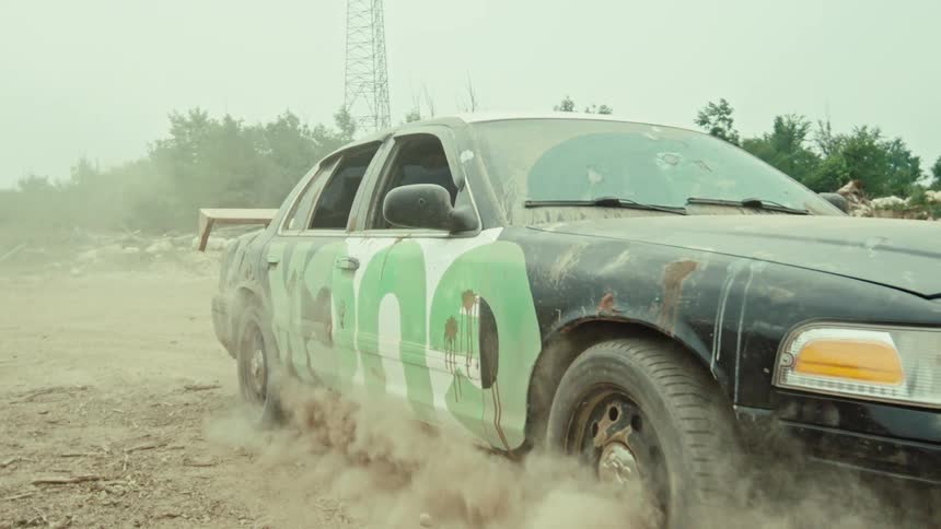 A custom-built demolition derby car, adorned with a green snake graphic, performs a burnout in a dusty junkyard in super slow motion