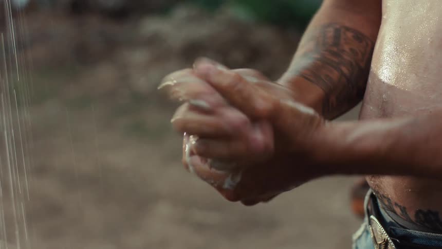 A shirtless tattooed man thoroughly washes his hands with soap and water in slow motion