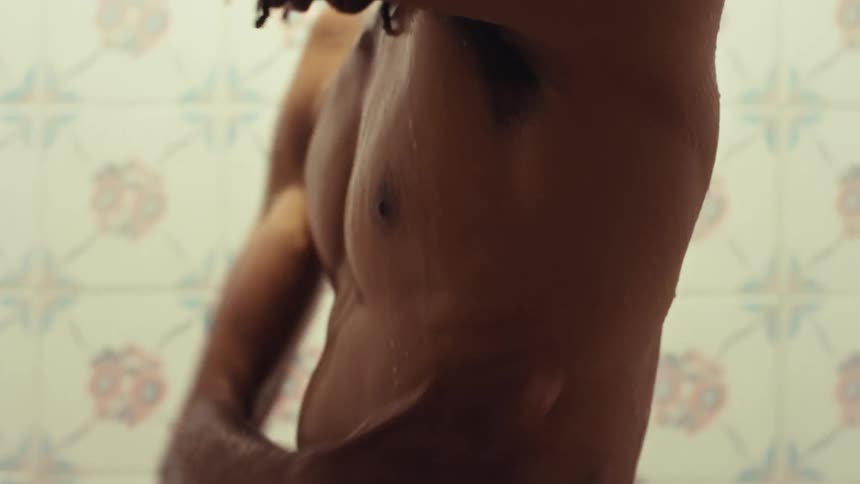 An intimate close-up shows a Black man with dreadlocks taking a shower, slowly washing his body under warm water