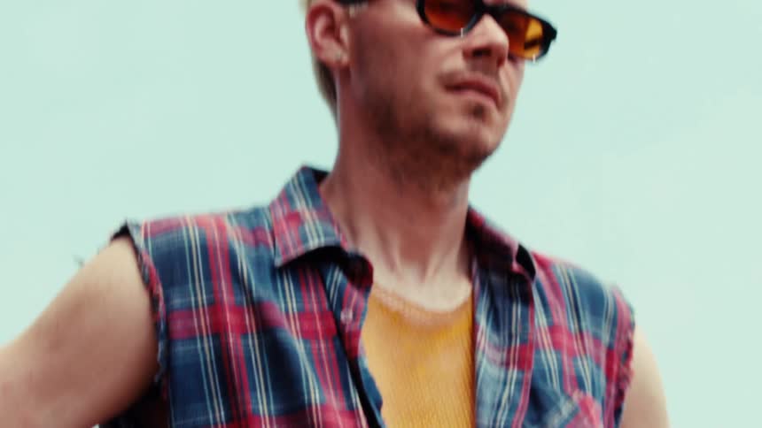A man with tattoos and blonde hair wearing a plaid shirt and sunglasses turns and walks away in slow motion
