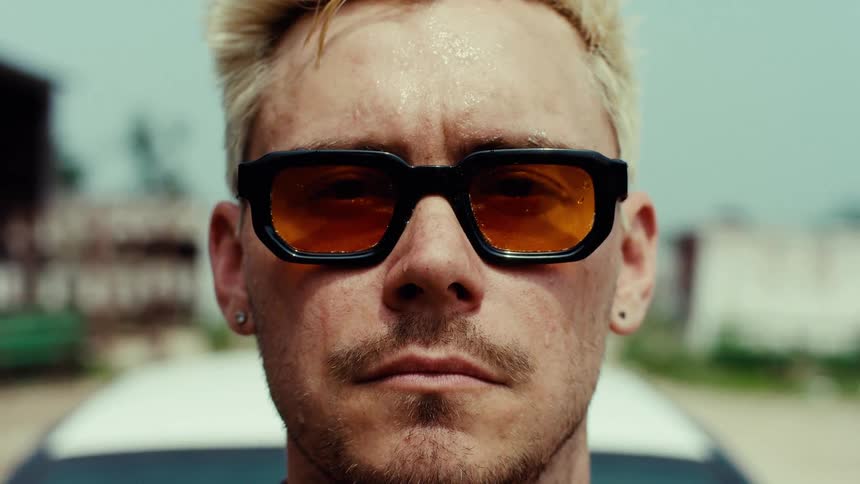 An intense close-up of a blonde man in orange sunglasses staring into the camera before quickly moving away