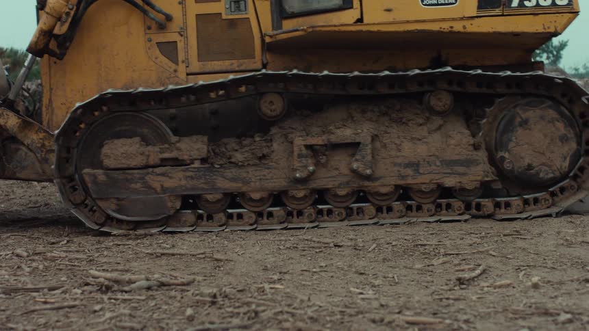 A low angle, slow pan shot of a muddy yellow bulldozer's tracks turning on a dirt field