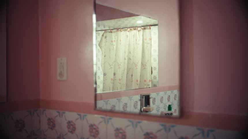 A person's reflection is seen in a pink-tiled bathroom mirror as they shower behind a floral curtain