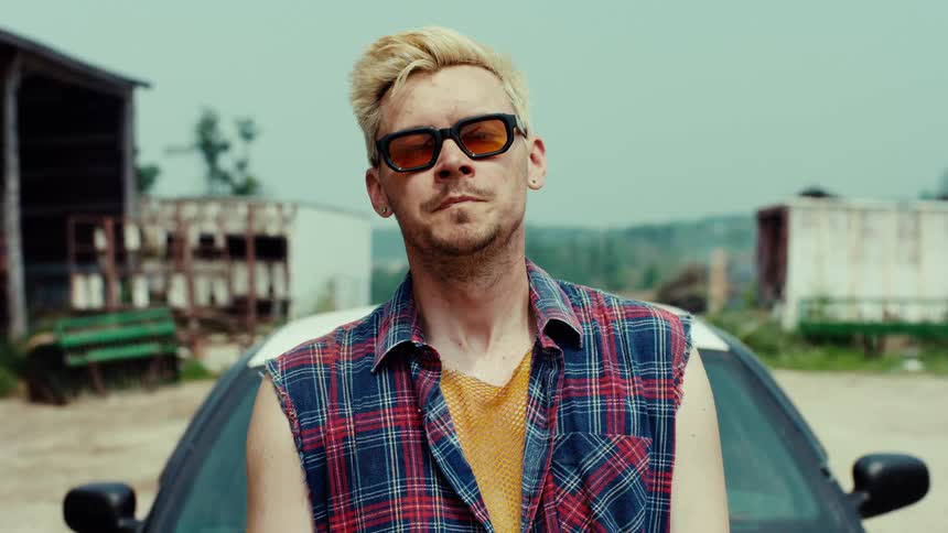 A tough-looking blonde man in sunglasses and a plaid shirt stares defiantly at the camera before walking away in a rural junkyard