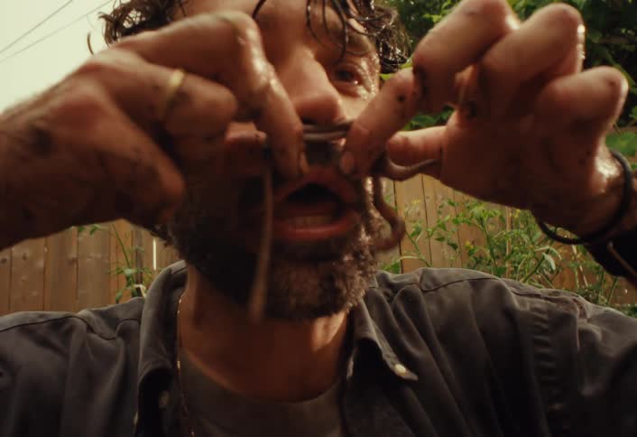 A disheveled man with dirty hands pulls apart a live earthworm and eats it in slow motion