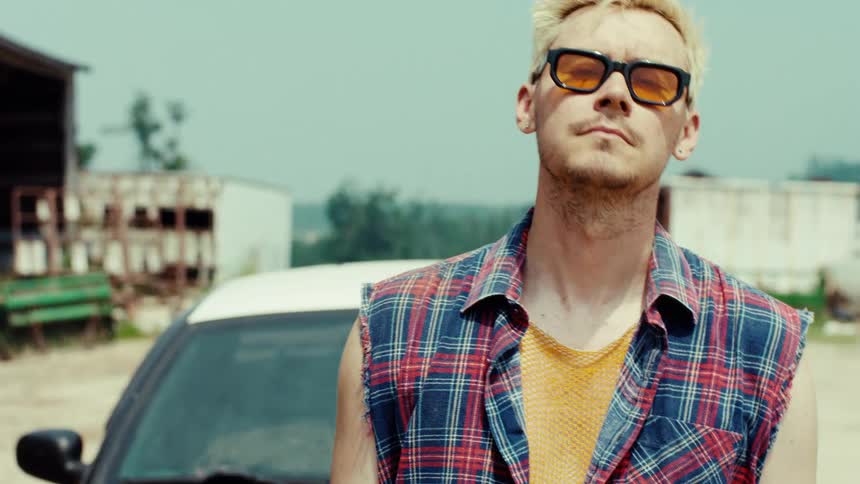 A confident young man with bleached blonde hair and sunglasses walks into focus and poses in front of a car in a rural setting