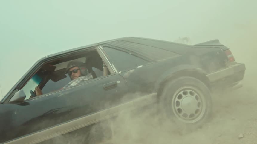 A man in a helmet drives a classic black muscle car through a massive cloud of dust in slow motion
