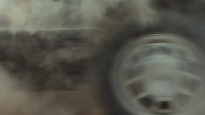 A low-angle slow-motion shot captures a classic black muscle car peeling out on a dirt road, its spinning tire kicking up a huge cloud of dust