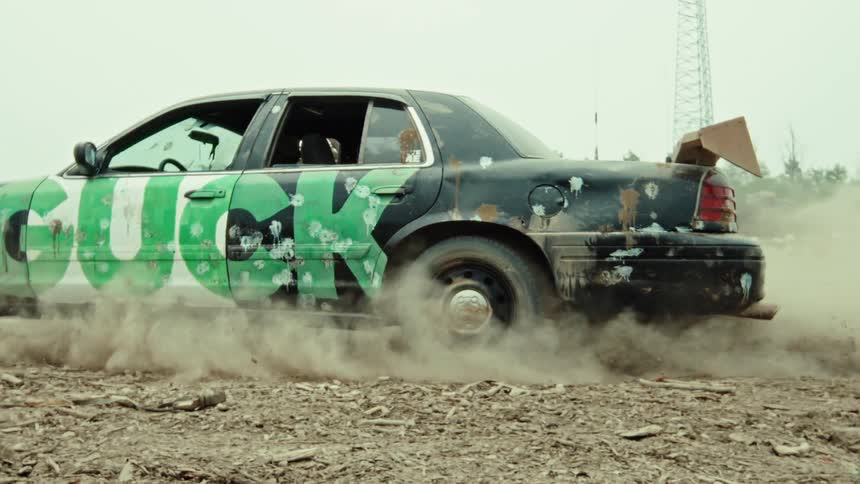 A heavily modified black car with green graffiti does a burnout in slow motion, kicking up a massive cloud of dust in a desolate, industrial wasteland