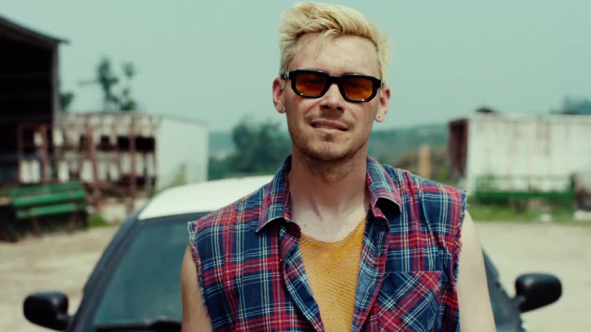A confident young man with bleached blonde hair and sunglasses poses in front of his car in a gritty rural setting