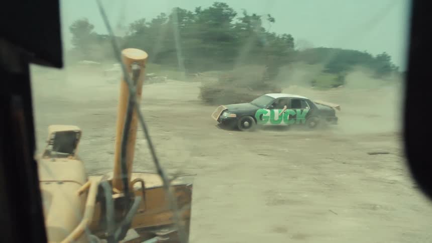 Point of view from inside a bulldozer as it slowly chases a demolition derby car through a dusty junkyard in slow motion