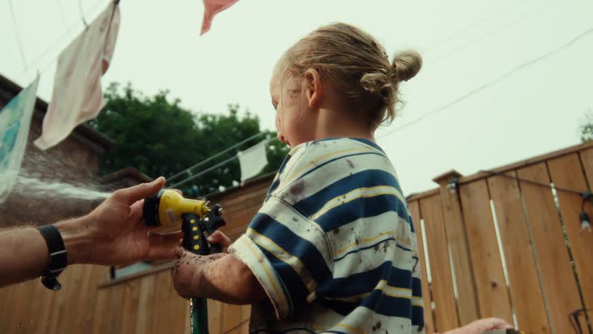 A low angle, slow-motion shot shows a muddy child spraying a garden hose with the help of a parent in the backyard