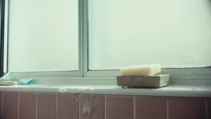 A soapy hand reaches up from out of frame to grab a bottle of body wash from a bathroom windowsill