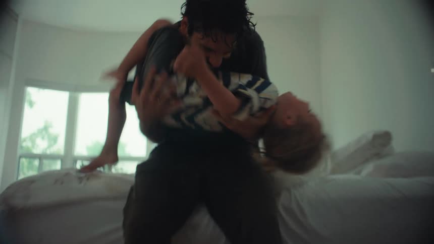 A dynamic low-angle shot races down a hallway into a bedroom where a father playfully swings his son around before falling onto the bed