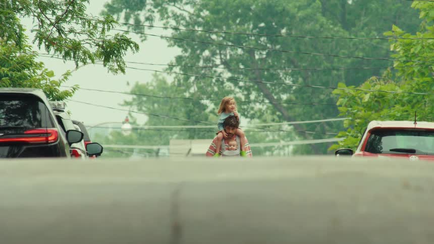 A father walks down the street carrying his baby in a carrier and his young daughter on his shoulders in slow motion