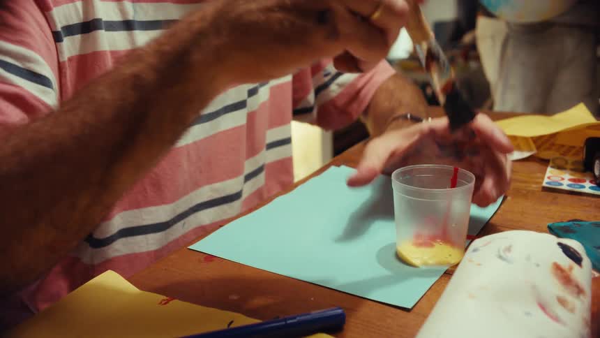 A male artist with paint on his face pours and mixes yellow and red paint in a cup with a brush at his art table