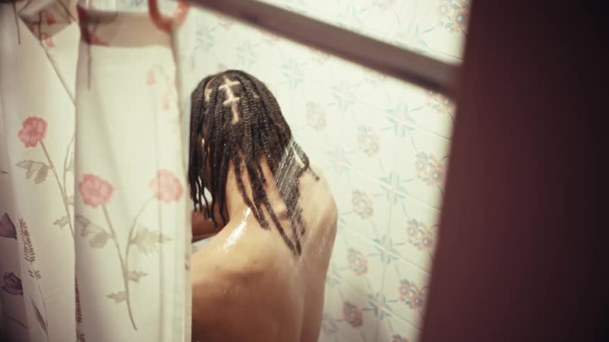 Over-the-shoulder shot of a person with braids washing their back with soap in the shower in slow motion