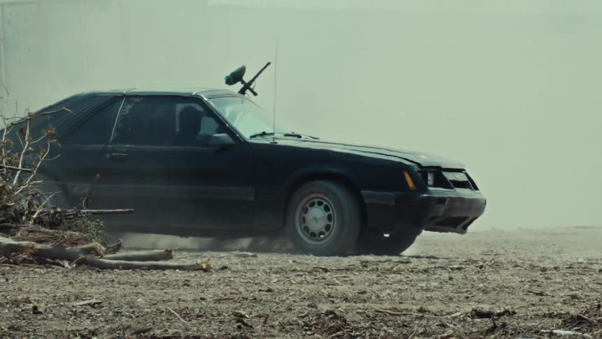A black sports car with a paintball gun mounted on it emerges from a cloud of dust in a gritty, wasteland setting