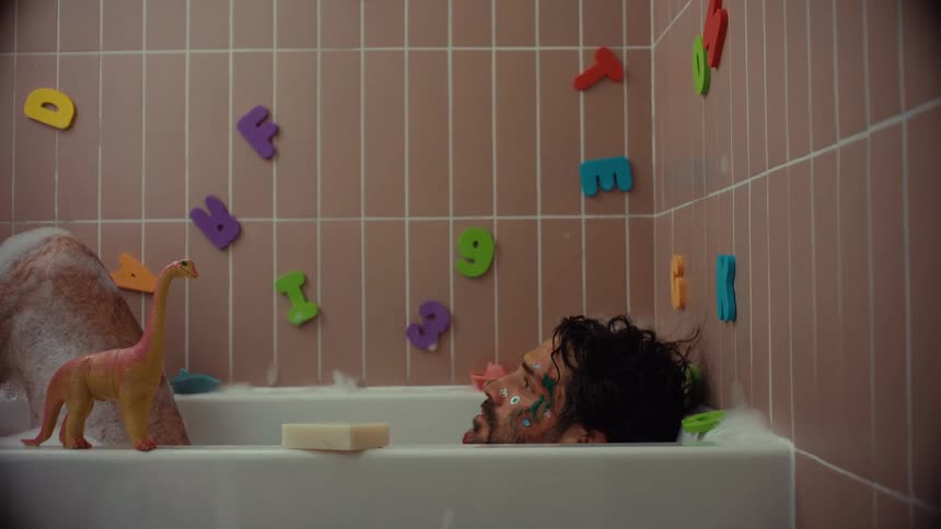 An exhausted father with paint on his face falls asleep in a bubble bath surrounded by his children's toys